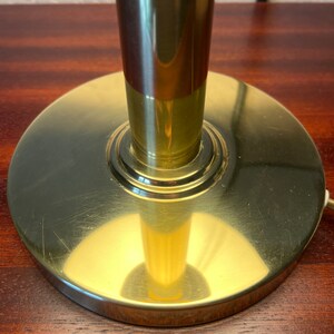 Puede incluir: Primer plano de una base de l&aacute;mpara de lat&oacute;n. La base es circular y plana, con un v&aacute;stago cil&iacute;ndrico. El lat&oacute;n tiene un acabado pulido con peque&ntilde;os ara&ntilde;azos. La base de la l&aacute;mpara est&aacute; sobre una superficie de madera oscura.