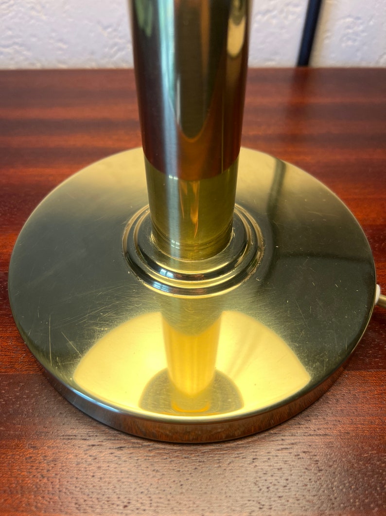 Puede incluir: Primer plano de una base de l&aacute;mpara de lat&oacute;n. La base es circular y plana, con un v&aacute;stago cil&iacute;ndrico. El lat&oacute;n tiene un acabado pulido con peque&ntilde;os ara&ntilde;azos. La base de la l&aacute;mpara est&aacute; sobre una superficie de madera oscura.