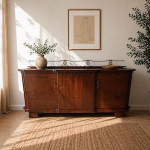 Peut inclure: Buffet en bois foncé avec une porte centrale et deux portes latérales. Au-dessus du buffet se trouve une barre métallique décorative avec trois motifs. Un croquis encadré est accroché au mur au-dessus du buffet. Un vase avec de la verdure est posé sur le buffet.