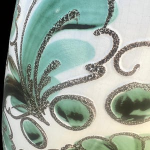 May include: A white ceramic vase with a teal and black floral design. The vase has a glossy finish and a slightly textured surface. The floral pattern features stylized leaves and oval shapes outlined in black.