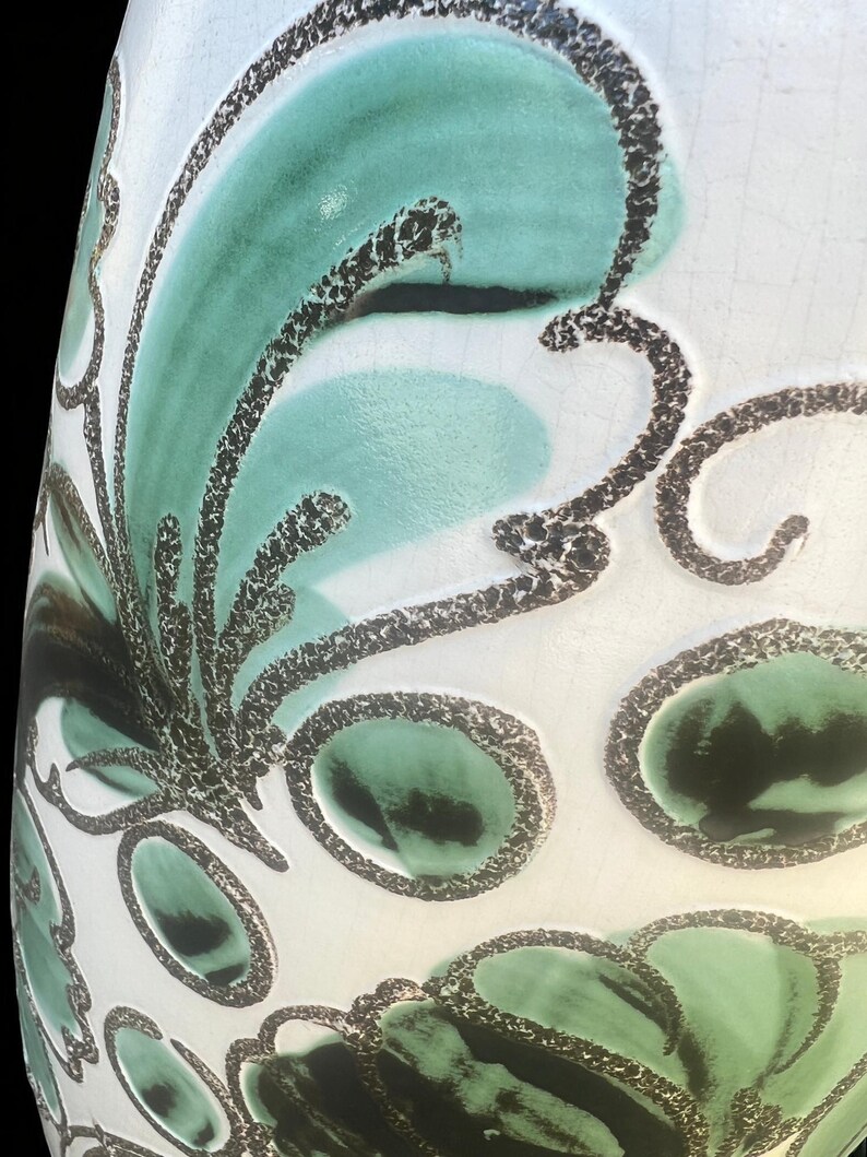 May include: A white ceramic vase with a teal and black floral design. The vase has a glossy finish and a slightly textured surface. The floral pattern features stylized leaves and oval shapes outlined in black.
