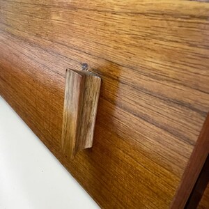 Puede incluir: Primer plano de un caj&oacute;n de madera con un tirador rectangular de madera. El caj&oacute;n es de madera marr&oacute;n claro con vetas visibles. El tirador es de un tono m&aacute;s claro y tiene un accesorio transparente similar al pl&aacute;stico.