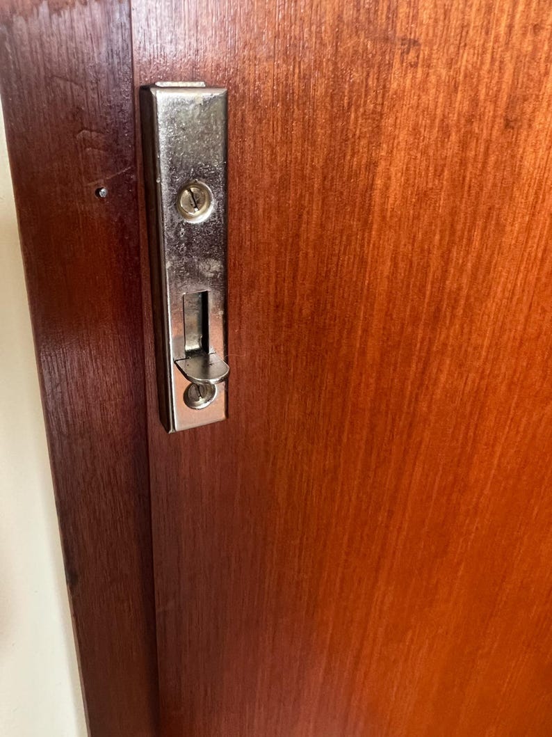 Puede incluir: Primer plano de una puerta de madera con una cerradura corredera de metal plateado brillante. La puerta es de color marr&oacute;n rojizo c&aacute;lido, con vetas de madera visibles. La cerradura tiene un ojo de cerradura y un mecanismo deslizante.