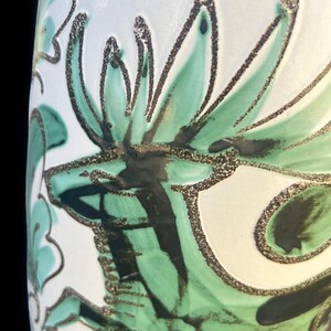 May include: Close-up of a white ceramic vase with a floral design. The design features green and black stylized leaves and petals with a textured, speckled outline. The vase has a glossy finish.