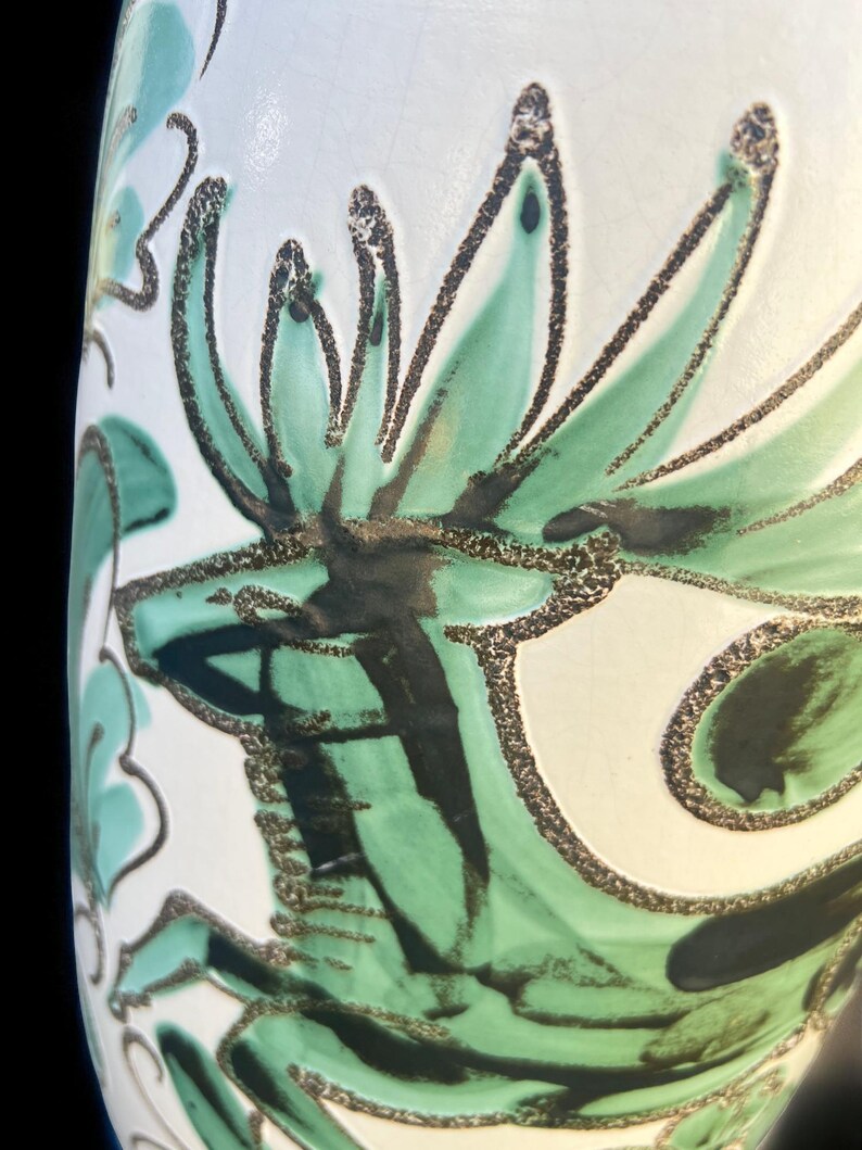 May include: Close-up of a white ceramic vase with a floral design. The design features green and black stylized leaves and petals with a textured, speckled outline. The vase has a glossy finish.