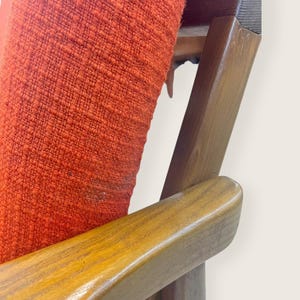 May include: Close-up of a mid-century modern chair. The chair features a vibrant orange fabric seat and backrest, contrasted by the warm tones of the wooden frame and armrests. The design showcases clean lines and a classic aesthetic, perfect for vintage furniture enthusiasts.