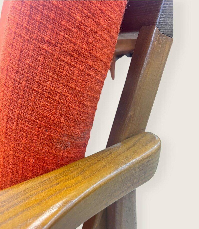 May include: Close-up of a mid-century modern chair. The chair features a vibrant orange fabric seat and backrest, contrasted by the warm tones of the wooden frame and armrests. The design showcases clean lines and a classic aesthetic, perfect for vintage furniture enthusiasts.