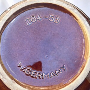 May include: Close-up of the base of a brown ceramic vessel. The base is a deep brown color with the embossed text "W. GERMANY" and the numbers "284-53". The rim is a lighter brown color.