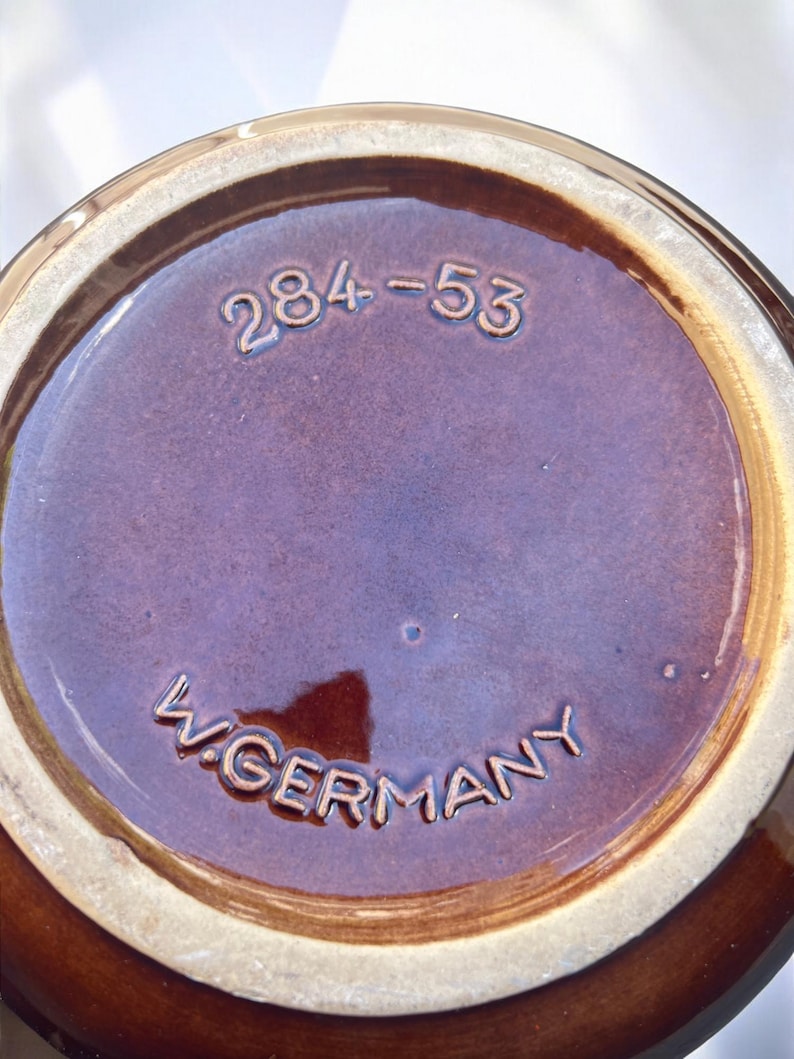 May include: Close-up of the base of a brown ceramic vessel. The base is a deep brown color with the embossed text "W. GERMANY" and the numbers "284-53". The rim is a lighter brown color.