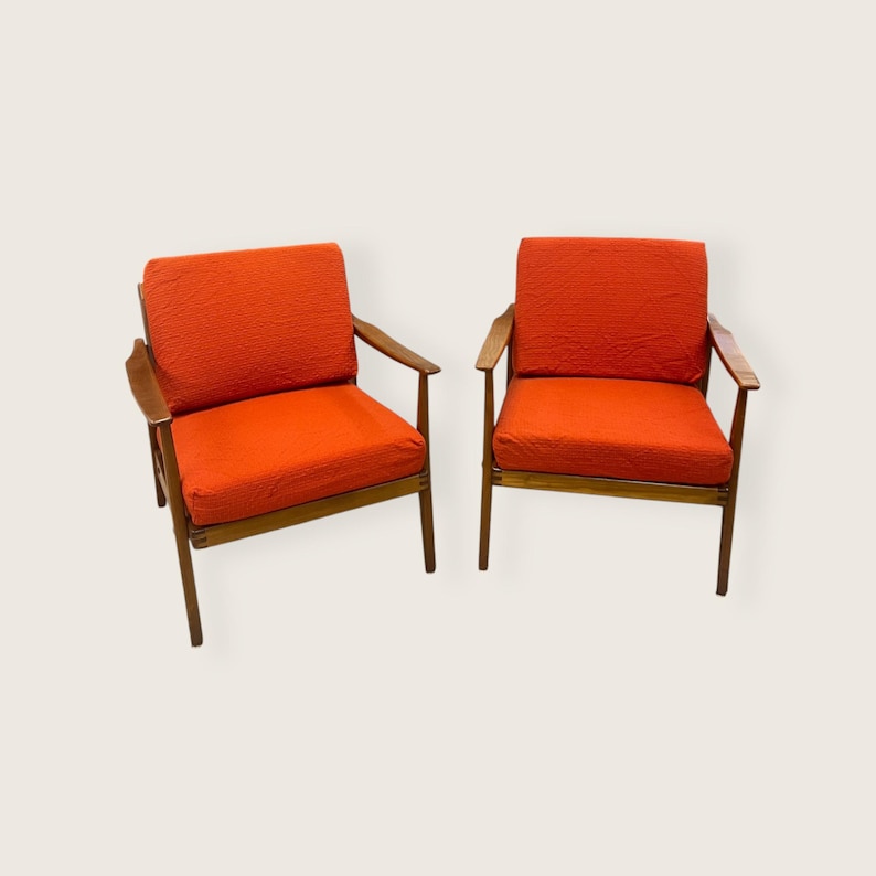 May include: Two mid-century modern armchairs with orange textured cushions and wooden frames. The chairs have a simple design with angled legs and armrests, perfect for a living room or lounge area.