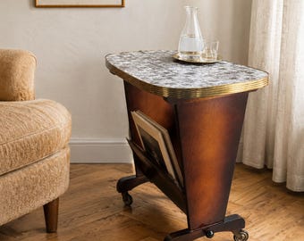 Puede incluir: Una mesa auxiliar móvil con acabado en madera oscura, superficie de mosaico y borde de metal color latón. La mesa cuenta con un revistero integrado que contiene dos libros y se apoya sobre cuatro ruedas pequeñas. Una jarra de cristal está encima.