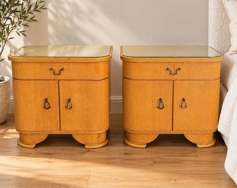Puede incluir: Un par de mesitas de noche de madera de color marrón claro, cada una con un cajón sobre dos puertas de armario con herrajes de metal oscuro y una superficie superior de cristal. Las mesitas tienen bases curvas y un acabado de madera.