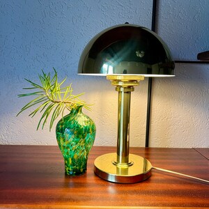 Puede incluir: Una l&aacute;mpara de escritorio de lat&oacute;n con una pantalla de c&uacute;pula verde oscuro y una base redonda. Junto a ella, un jarr&oacute;n de vidrio moteado verde y amarillo contiene una ramita de vegetaci&oacute;n. La l&aacute;mpara est&aacute; sobre una superficie de madera oscura.