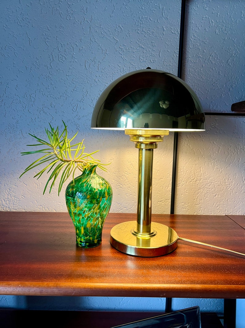 Puede incluir: Una l&aacute;mpara de escritorio de lat&oacute;n con una pantalla de c&uacute;pula verde oscuro y una base redonda. Junto a ella, un jarr&oacute;n de vidrio moteado verde y amarillo contiene una ramita de vegetaci&oacute;n. La l&aacute;mpara est&aacute; sobre una superficie de madera oscura.