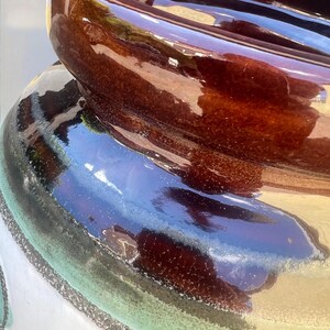 May include: Close-up of a ceramic pot with a glossy, brown rim and a dark brown interior. The pot's exterior features a gradient of colors, including white, green, and blue, with decorative black accents. The pot has a wide opening.