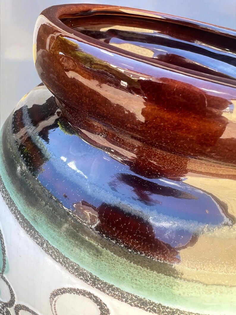 May include: Close-up of a ceramic pot with a glossy, brown rim and a dark brown interior. The pot's exterior features a gradient of colors, including white, green, and blue, with decorative black accents. The pot has a wide opening.