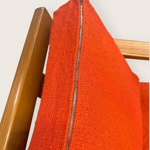 May include: Close-up of a vibrant orange fabric with a silver zipper. The textured material is likely part of a garment, possibly a dress or skirt, hanging on a wooden hanger. The bright color and metallic zipper create a striking contrast.