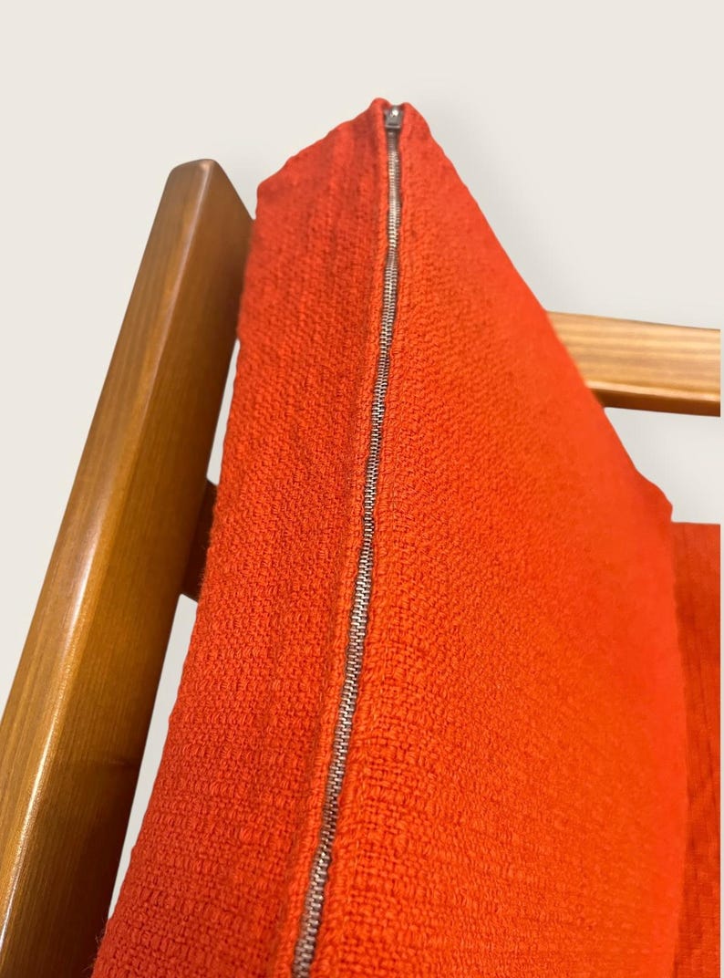 May include: Close-up of a vibrant orange fabric with a silver zipper. The textured material is likely part of a garment, possibly a dress or skirt, hanging on a wooden hanger. The bright color and metallic zipper create a striking contrast.