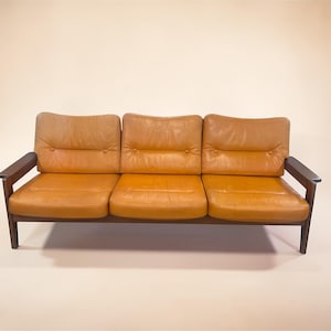 May include: A three-seat sofa with a mid-century modern design. The sofa features a warm, tan leather upholstery and dark wooden armrests and frame. The cushions are tufted with button detailing.