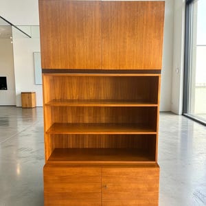 Puede incluir: Un mueble alto de madera en color marr&oacute;n claro. Cuenta con puertas superiores, estanter&iacute;as abiertas y puertas inferiores. El dise&ntilde;o incorpora una base y molduras de color marr&oacute;n oscuro, contrastando con la madera m&aacute;s clara. El mueble es adecuado para el almacenamiento, aprox. 180 cm de altura.