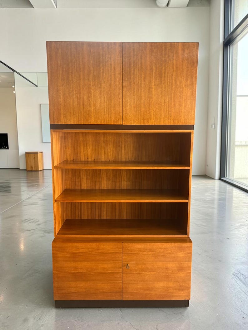 Puede incluir: Un mueble alto de madera en color marr&oacute;n claro. Cuenta con puertas superiores, estanter&iacute;as abiertas y puertas inferiores. El dise&ntilde;o incorpora una base y molduras de color marr&oacute;n oscuro, contrastando con la madera m&aacute;s clara. El mueble es adecuado para el almacenamiento, aprox. 180 cm de altura.