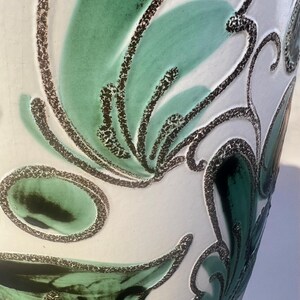 May include: Close-up of a white ceramic vase with a textured surface. The vase features a design of green and black abstract floral patterns. The green patterns have a speckled texture, and the black outlines are thick and dotted.