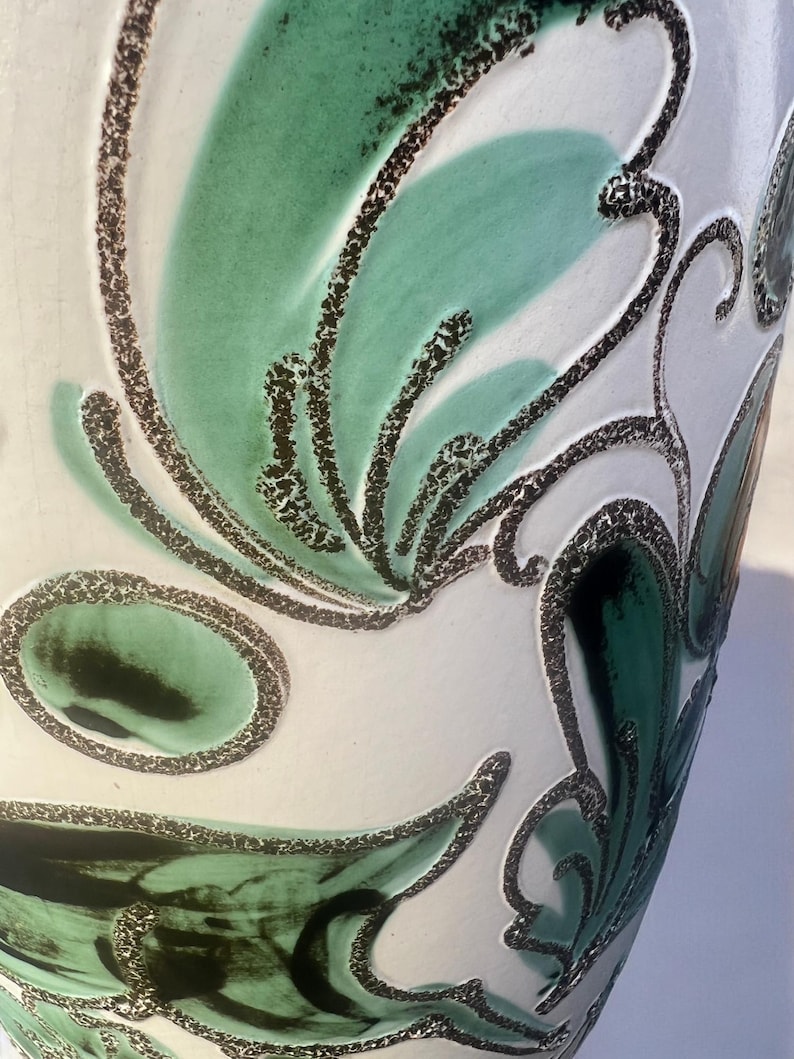 May include: Close-up of a white ceramic vase with a textured surface. The vase features a design of green and black abstract floral patterns. The green patterns have a speckled texture, and the black outlines are thick and dotted.