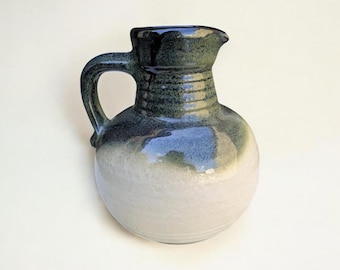 May include: A handmade ceramic pitcher with a rounded body and a handle. The pitcher is glazed in shades of green, blue, and white, with a dark green rim and handle. The design is simple and rustic, suitable for serving water or other beverages.