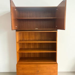 Puede incluir: Una estanter&iacute;a de madera con un acabado marr&oacute;n rojizo c&aacute;lido. La secci&oacute;n superior tiene dos puertas que se abren hacia afuera, revelando estantes. La secci&oacute;n media tiene estantes abiertos y la parte inferior tiene dos puertas. Estilo moderno de mediados de siglo.