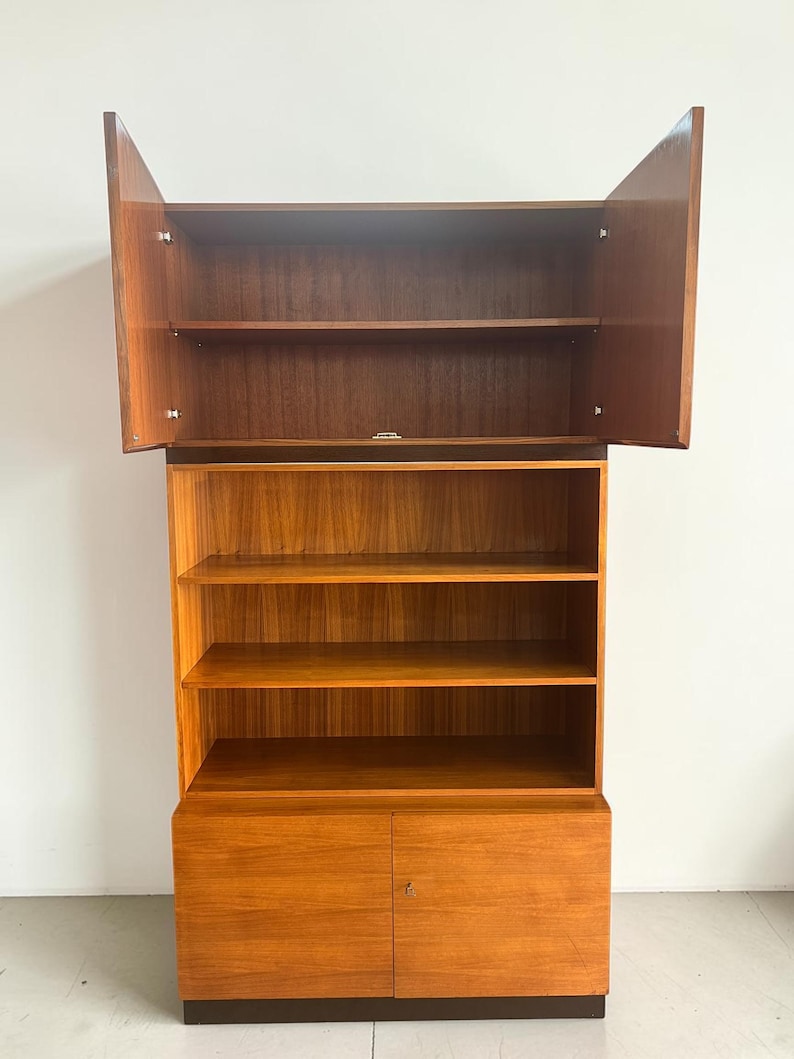 Puede incluir: Una estanter&iacute;a de madera con un acabado marr&oacute;n rojizo c&aacute;lido. La secci&oacute;n superior tiene dos puertas que se abren hacia afuera, revelando estantes. La secci&oacute;n media tiene estantes abiertos y la parte inferior tiene dos puertas. Estilo moderno de mediados de siglo.