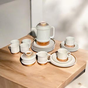 Puede incluir: Un juego de té de porcelana blanca con detalles dorados. El juego incluye una tetera, una jarra de crema, un azucarero y seis tazas y platillos.