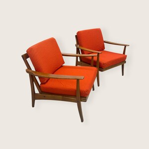 May include: Two mid-century modern armchairs with orange textured cushions and wooden frames. The chairs have a low profile with angled legs and armrests, showcasing a classic design. The cushions are a vibrant orange, contrasting with the warm wood tones.