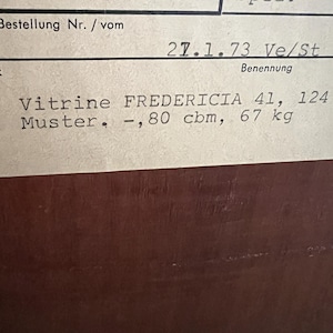 Puede incluir: Un primer plano de una etiqueta blanca con texto negro. La etiqueta dice "Komm.-Nr. 51/212", "Bestellung Nr. / vom 27.1.73 Ve/St", "Benennung", "Vitrine FREDERICIA 41, 124", "Muster. -,80 cbm, 67 kg".
