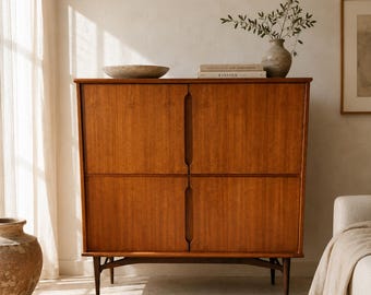Puede incluir: Un armario rectangular alto de madera con un acabado marrón cálido, que presenta cuatro puertas con tiradores verticales empotrados. El mueble se apoya sobre cuatro patas cónicas y tiene una superficie superior lisa con un cuenco de piedra.