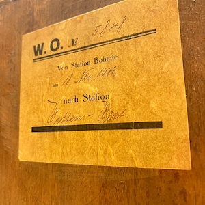 May include: A vintage luggage tag with the text "W.O. Nr. 5848 Von Station Bohmte am 18 Nov 1914 nach Station" written in black ink on a yellowed paper background.