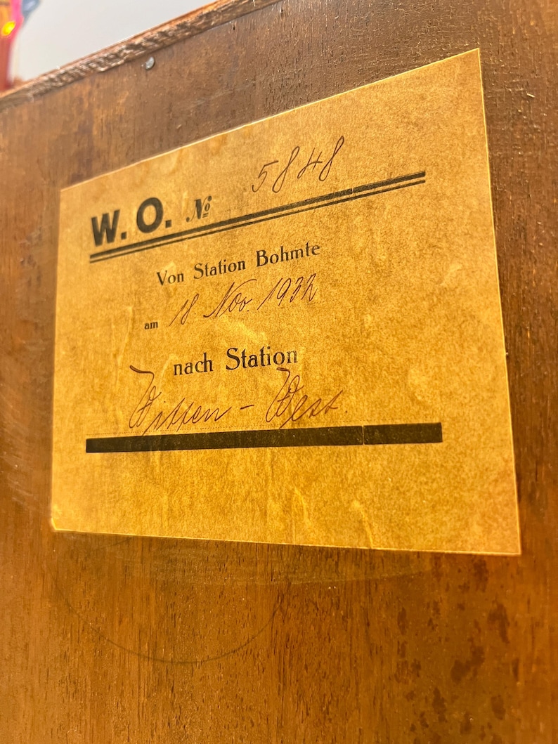 May include: A vintage luggage tag with the text "W.O. Nr. 5848 Von Station Bohmte am 18 Nov 1914 nach Station" written in black ink on a yellowed paper background.