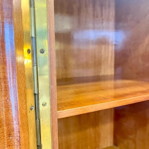 May include: Close-up of a wooden cabinet with a glass door and brass hinges. The cabinet has two shelves and is made of a light wood.