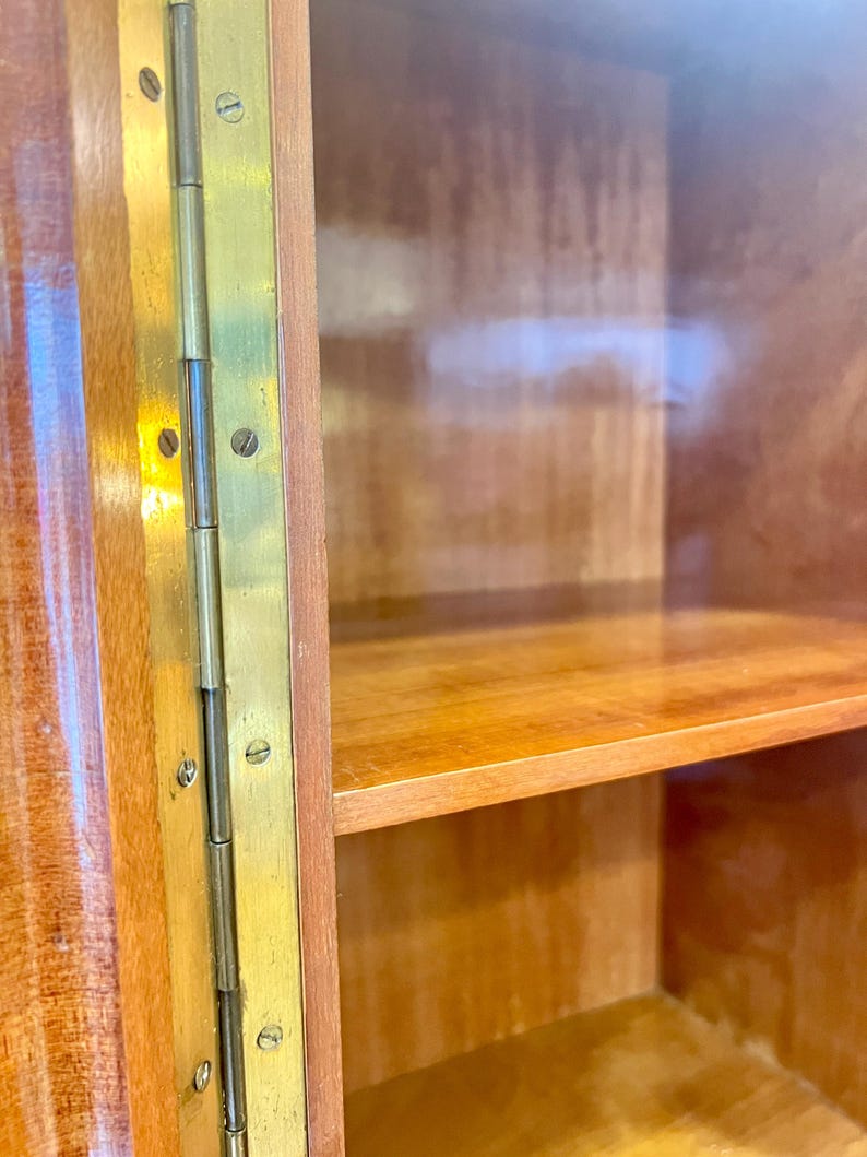 May include: Close-up of a wooden cabinet with a glass door and brass hinges. The cabinet has two shelves and is made of a light wood.