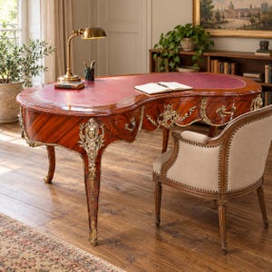 May include: A kidney-shaped wooden desk with a red leather top, ornate gold-toned metal mounts, and curved legs. The desk features multiple drawers with metal pulls, a brass desk lamp, a pen holder, and an open book resting on the surface.