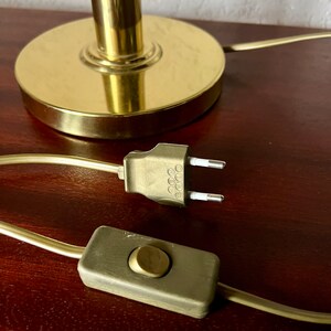 Puede incluir: Primer plano de una base de l&aacute;mpara dorada, cable de alimentaci&oacute;n y enchufe. La base de la l&aacute;mpara es circular con un v&aacute;stago cil&iacute;ndrico. El cable de alimentaci&oacute;n tiene un interruptor y un enchufe de dos clavijas.