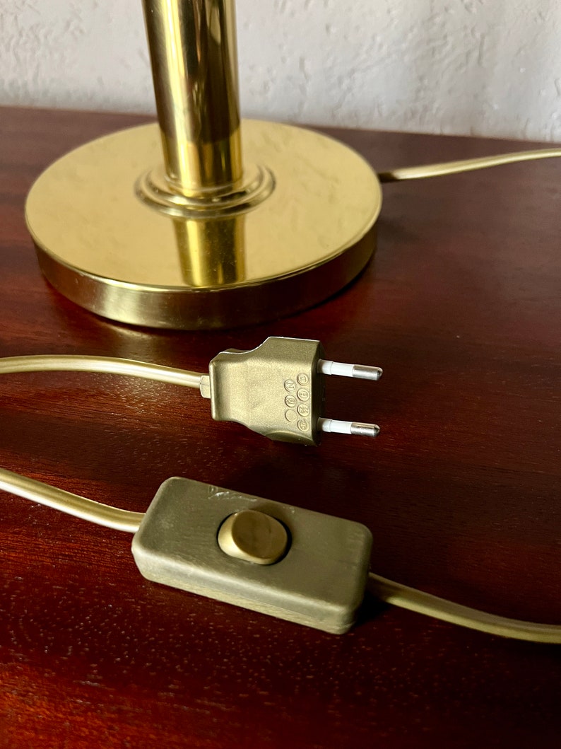 Puede incluir: Primer plano de una base de l&aacute;mpara dorada, cable de alimentaci&oacute;n y enchufe. La base de la l&aacute;mpara es circular con un v&aacute;stago cil&iacute;ndrico. El cable de alimentaci&oacute;n tiene un interruptor y un enchufe de dos clavijas.