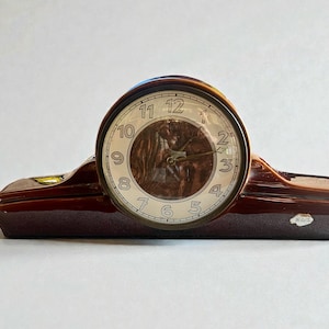 May include: A brown ceramic mantel clock with a cream-colored clock face and black hands. The clock has a curved, rectangular base and a rounded top. The clock face features black numerals and a textured, brown center.