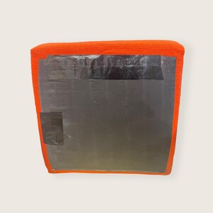May include: An orange, square-shaped cushion with a textured fabric. The cushion is covered with a large, dark gray sheet, secured with black tape. A smaller piece of black tape is on the lower left corner.