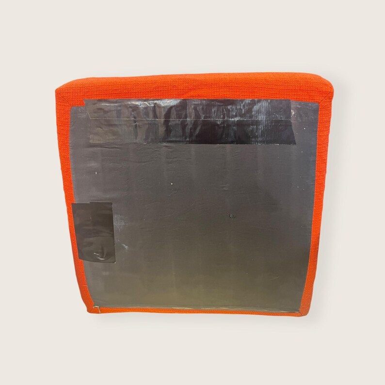 May include: An orange, square-shaped cushion with a textured fabric. The cushion is covered with a large, dark gray sheet, secured with black tape. A smaller piece of black tape is on the lower left corner.