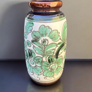 May include: A ceramic vase with a white base, adorned with green floral patterns. The vase has a brown rim and a brown base. The floral design is outlined in a glittery silver color. The vase is a decorative item.