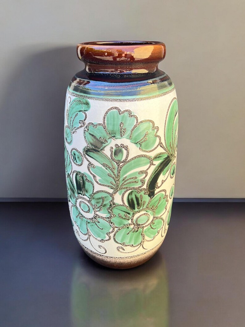 May include: A ceramic vase with a white base, adorned with green floral patterns. The vase has a brown rim and a brown base. The floral design is outlined in a glittery silver color. The vase is a decorative item.