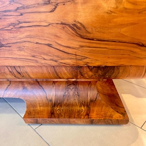 May include: A close-up of a wooden table base with a curved design. The wood has a rich brown color with dark brown grain patterns.