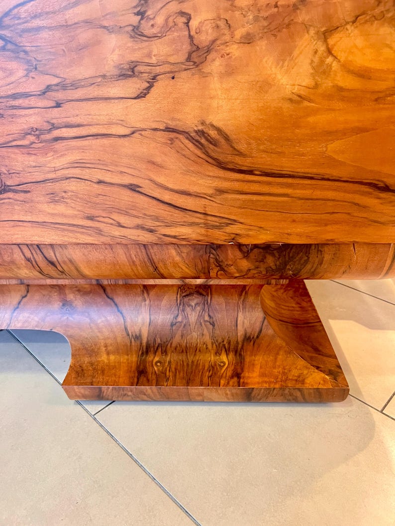 May include: A close-up of a wooden table base with a curved design. The wood has a rich brown color with dark brown grain patterns.