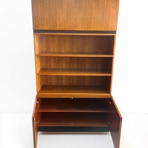 Puede incluir: Un mueble de madera de estilo moderno de mediados de siglo. El mueble tiene una estructura alta y vertical con almacenamiento cerrado en la parte superior e inferior, y estanter&iacute;as abiertas en el medio. La madera tiene un tono c&aacute;lido y natural, y el mueble est&aacute; sobre un fondo blanco.