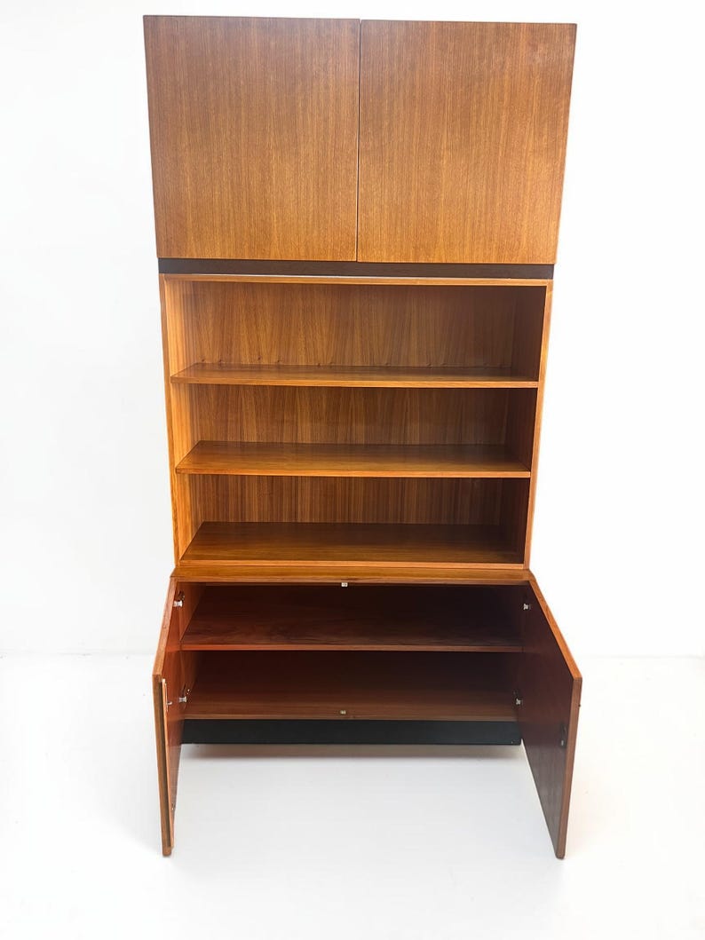 Puede incluir: Un mueble de madera de estilo moderno de mediados de siglo. El mueble tiene una estructura alta y vertical con almacenamiento cerrado en la parte superior e inferior, y estanter&iacute;as abiertas en el medio. La madera tiene un tono c&aacute;lido y natural, y el mueble est&aacute; sobre un fondo blanco.